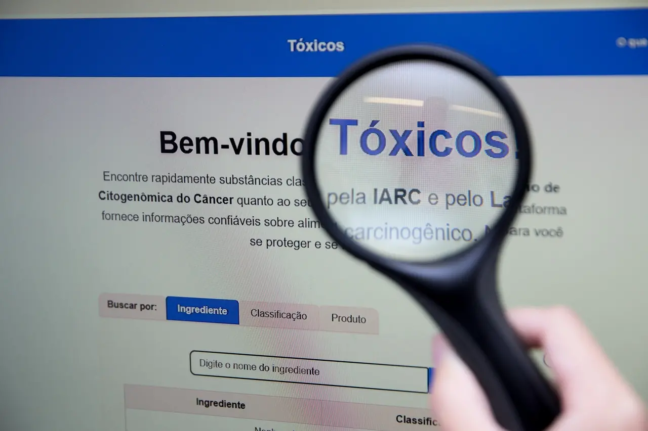 Imagem mostra mão segurando uma lupa em frente à tela de um computador que exibe a página inicial da plataforma digital Tóxicos. Por meio da lente da lupa, lê-se em destaque a palavra Tóxicos. Abaixo, na interface do site, aparecem botões de busca por Ingrediente, Classificação e Produto, além de uma barra de pesquisa com o texto Digite o nome do ingrediente.