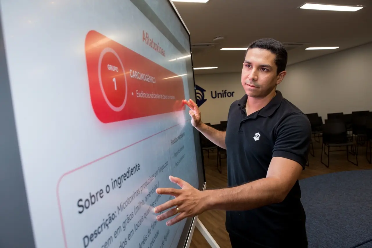 Pedro Pinheiro, coordenador tecnológico na Unifor, interage com um ecrã táctil de grande dimensão no laboratório. O ecrã apresenta informações detalhadas sobre a substância Aflatoxinas, classificando-a como GRUPO 1, CARCINOGÊNICO, com Evidências suficientes de cancro em humanos. O texto descreve as aflatoxinas como micotoxinas produzidas por fungos encontrados em grãos como o milho e o amendoim, particularmente em climas quentes. Pinheiro está a apontar para o ecrã, com o logótipo da Unifor visível ao fundo, ilustrando o trabalho de desenvolvimento e curadoria de dados da plataforma Tóxicos.