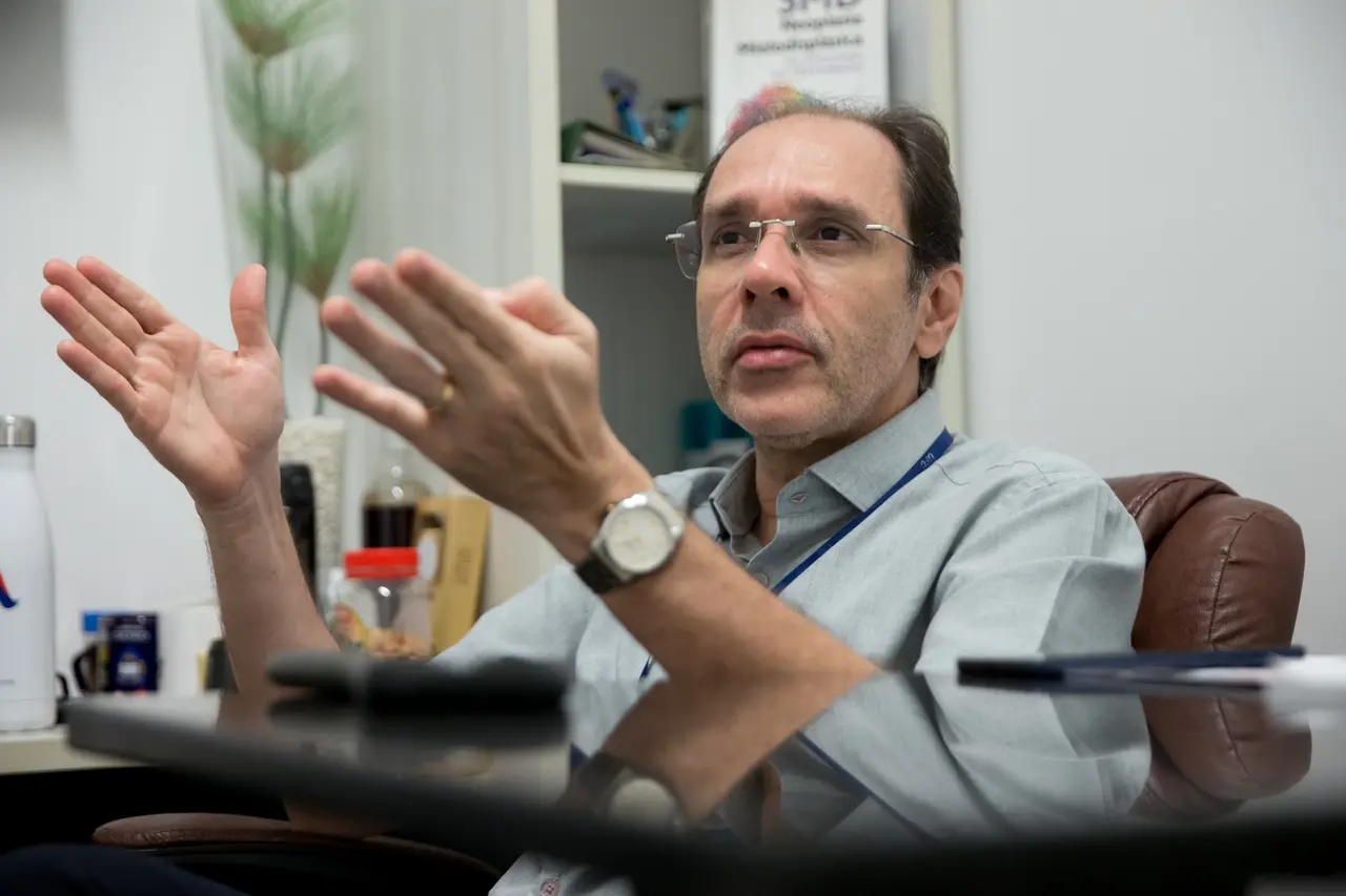Imagem mostra o médico hematologista e professor Ronald Feitosa Pinheiro, de óculos e camisa social cinza, sentado em uma cadeira de escritório de couro marrom, por trás de uma mesa escura e reflexiva. Ele gesticula com as duas mãos espalmadas enquanto fala, demonstrando entusiasmo. Ao fundo, de forma levemente desfocada, avista-se parte de uma estante com livros e objetos de escritório.