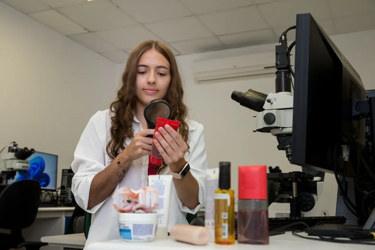 Imagem mostra a estudante de Biologia da UFC Sarah Cristina Carneiro, uma jovem de cabelos longos e ondulados, vestindo um jaleco branco, que está em um laboratório de pesquisa. Ela segura uma lupa com a mão direita para examinar o rótulo de uma embalagem plástica vermelha de cosmético, que segura com a mão esquerda. À sua frente, sobre a bancada branca, há outros produtos de higiene e beleza, além de um microscópio acoplado a um monitor de computador no lado direito da imagem.