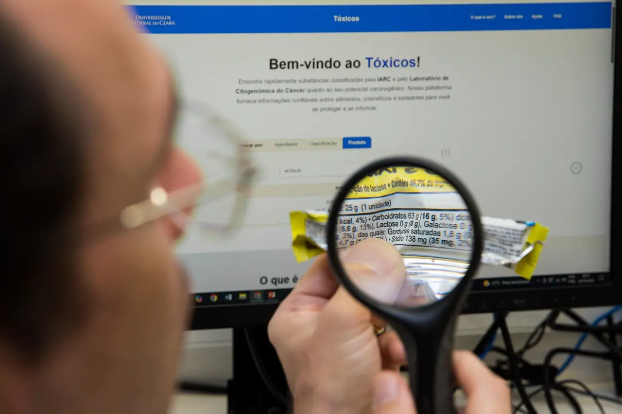 Em primeiro plano, no lado esquerdo e de perfil, a cabeça de um homem usando óculos de grau é vista parcialmente. Ele observa a tela de um computador ao fundo, que exibe o site da plataforma Tóxicos. Entre ele e o monitor, a sua mão direita segura uma lupa focada sobre o rótulo nutricional de uma barra de chocolate embalada em papel amarelo e aberto. Através da lente da lupa, as informações de ingredientes e a tabela nutricional do produto aparecem ampliadas e nítidas.