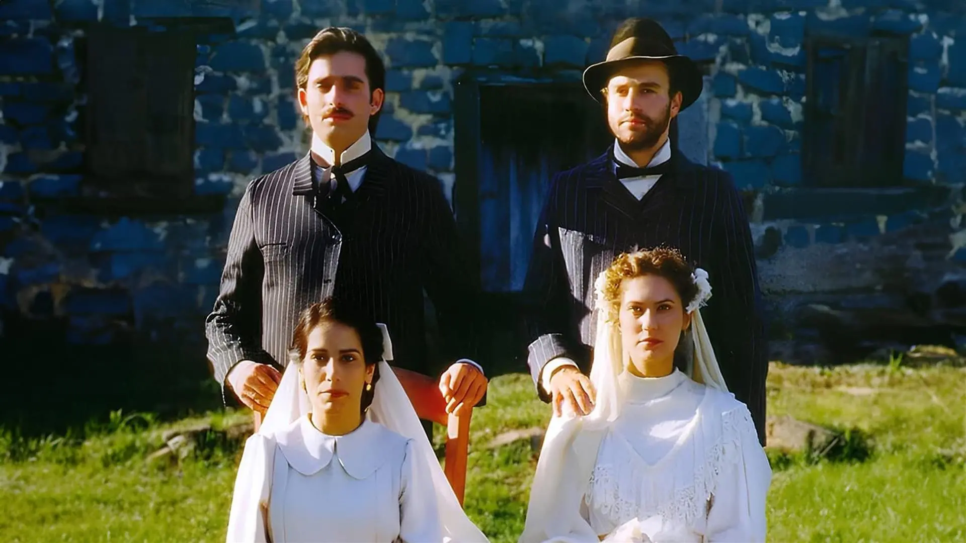 Cena do filme 'O Quatrilho' apresenta dois casais vestidos com trajes de época posando sérios em um ambiente externo, com as duas mulheres sentadas à frente usando vestidos e véus brancos. Atrás delas, os dois homens vestem ternos escuros riscados e golas altas, posicionados diante de uma parede de pedra rústica azulada sob a luz do dia.