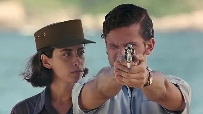 Cena do filme 'O Que É Isso, Companheiro?' mostra um homem em primeiro plano apontando firmemente um revólver em direção à câmera com as duas mãos, mantendo um dos olhos fechados para mirar. Ao fundo, uma mulher com quepe militar observa a ação com semblante sério, contra um cenário desfocado que sugere uma paisagem litorânea.