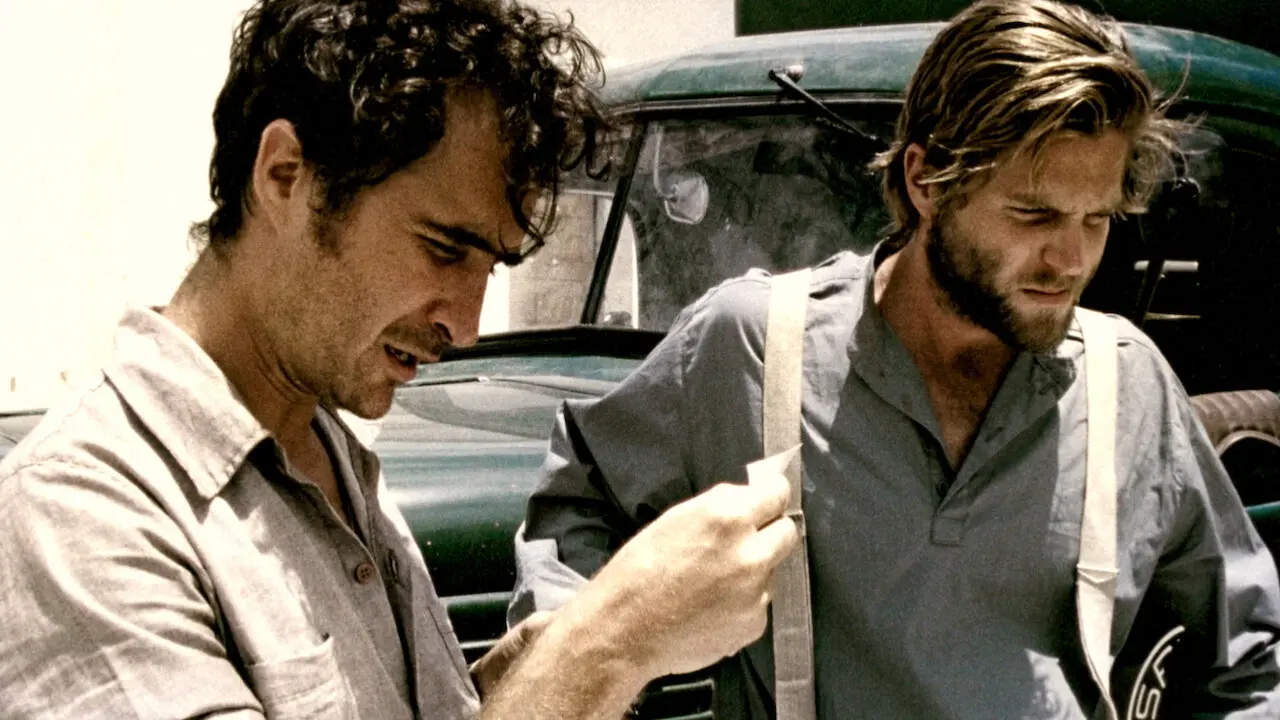 Cena do filme 'Cinema, Aspirinas e Urubus' retrata dois homens em primeiro plano sob a luz forte do sol, com o homem da esquerda segurando e analisando um pequeno papel branco enquanto fala. O outro homem, vestindo camisa de botões com suspensórios claros, observa a ação com semblante pensativo diante de um caminhão antigo de cor verde-escura ao fundo.