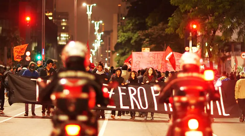 Manifestantes marcham por uma grande avenida urbana à noite segurando uma longa faixa preta enquanto são observados em primeiro plano por policiais em motocicletas com luzes acesas. O cenário ao fundo traz a iluminação pública da via e outros manifestantes com bandeiras, compondo uma cena de protesto em movimento sob a perspectiva dos motociclistas.