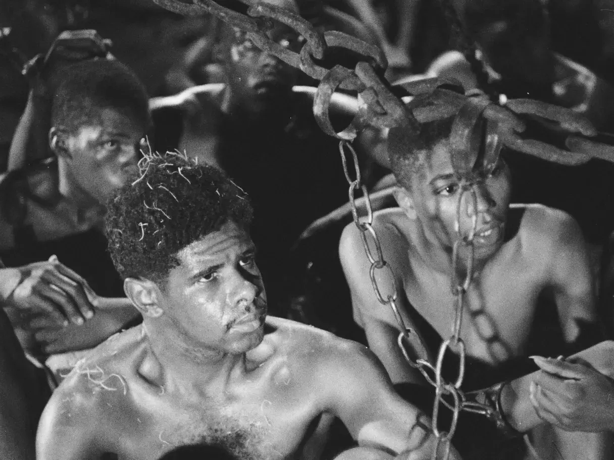 Homens negros com os torsos nus aparecem confinados em um ambiente escuro, com expressões marcadas por tensão e exaustão. Grossas correntes de ferro cruzam o primeiro plano e algemam seus pulsos, compondo uma cena dramática e de forte impacto em preto e branco.