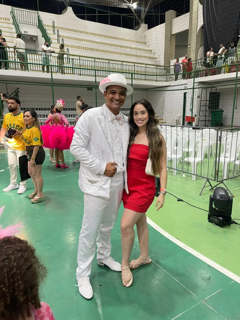 Na imagem, foto vertical de corpo inteiro de um casal sorrindo em uma quadra esportiva verde com linhas brancas. À esquerda, o homem veste um traje de noivo de quadrilha junina completamente branco, composto por paletó decorado com pequenos brilhos na gola e nos punhos, camisa social texturizada com gravata borboleta, calça e sapatos sociais brancos. Ele usa um chapéu de aba branco com uma flor rosa presa na lateral. À direita, a mulher tem cabelos longos e ondulados e usa um vestido curto e justo sem alças na cor vermelha, sandálias rasteiras de tiras claras e carrega uma pequena bolsa branca no ombro. Ao fundo, vê-se a movimentação do ginásio: pessoas circulando com camisas amarelas do Brasil e trajes juninos rosa, fileiras de cadeiras de plástico brancas organizadas atrás de grades de proteção e, mais acima, uma arquibancada de concreto com corrimãos verdes onde algumas pessoas observam o evento.