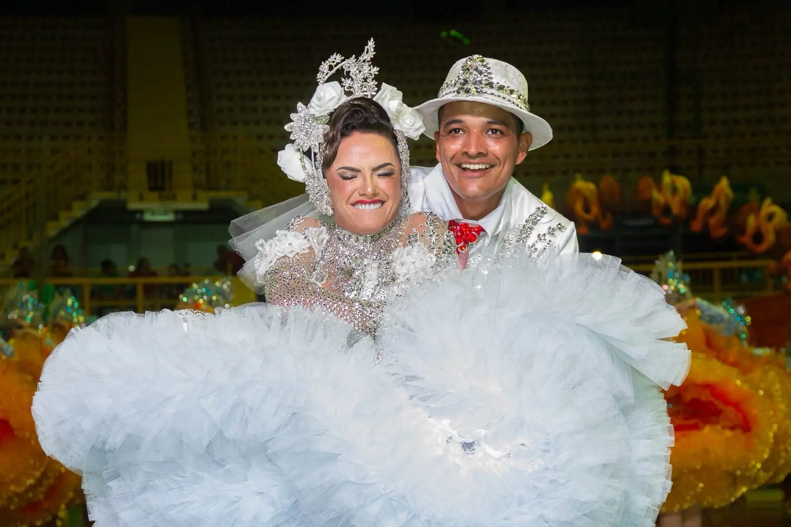 Na imagem, uma fotografia de corpo inteiro e plano médio de um casal de noivos de quadrilha junina posando em um ginásio. A noiva, em primeiro plano, tem uma expressão dramática de choro e riso, vestindo um vestido branco de tule extremamente volumoso e ornamentado com cristais e glitter, e um arranjo de cabeça floral prateado e branco. O noivo, posicionado atrás dela e sorrindo amplamente, veste um terno branco com glitter e uma gravata vermelha, e um chapéu fedora branco decorado com cristais. Eles estão sobre uma quadra de ginásio polida. Ao fundo, as arquibancadas mostram banners e logotipos distintos do 'CEARÁ SC' e da mascote 'VOZÃO' da equipe, juntamente com outros dançarinos de quadrilha desfocados. A iluminação é nítida e brilhante, destacando os detalhes complexos dos trajes.