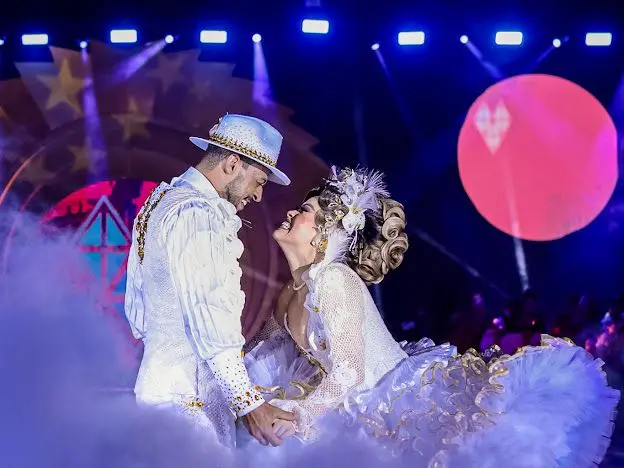 Na imagem, fotografia de um casal de dançarinos de festa junina (quadrilha) sorrindo e olhando fixamente um para o outro em uma pose romântica no palco. À esquerda, o homem veste um traje de noivo branco com detalhes dourados e um chapéu de aba curto decorado. À direita, a mulher usa um vestido de noiva rodado e volumoso, também branco com bordas douradas, além de um penteado elaborado com arranjo de flores e penas na cabeça. Eles estão de mãos dadas, cercados por um efeito de fumaça branca na altura da cintura. O fundo é escuro, iluminado por refletores azuis no topo, um painel circular com estrelas à esquerda e uma grande lua cheia cenográfica em tom rosa-avermelhado à direita.