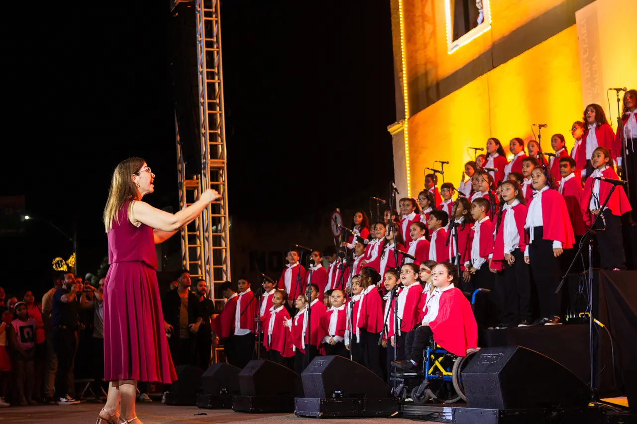 Uma maestrina de vestido magenta rege, com os braços erguidos, um coral infantil posicionado em arquibancadas sob a iluminação calorosa de um edifício histórico à noite. As crianças vestem uniformes com capas vermelhas e camisas brancas, destacando uma delas em uma cadeira de rodas na primeira fila enquanto cantam com entusiasmo.