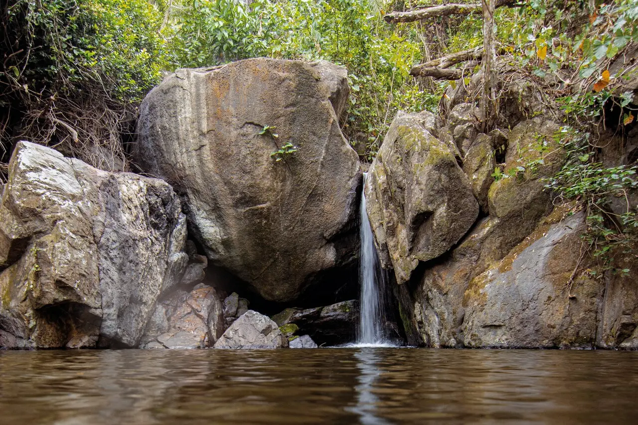 Pequena queda d'água fluindo entre grandes rochas e paredões de pedra cercados por vegetação densa na Serra da Aratanha, no Ceará. O cenário natural ilustra uma das formações geográficas do estado classificadas como montanhas pelo IBGE.