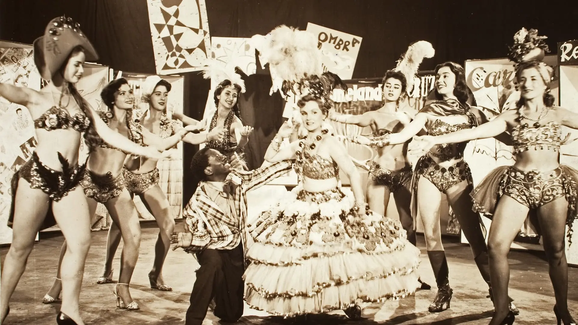 Artistas, incluindo o humorista Grande Otelo de blusa xadrez e ajoelhado, performam em um palco decorado, destacando uma dançarina com um vestido de babados volumoso no centro. O grupo de passistas com trajes festivos e adereços de plumas compõe a cena antiga em tons de sépia, evocando uma atmosfera vibrante de dança e espetáculo musical.
