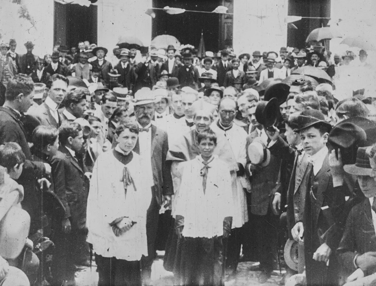 Imagem de reprodução de filme histórico de 1910 mostra pessoas que se reúnem em frente a uma igreja, destacando dois jovens coroinhas em trajes brancos no centro enquanto uma multidão de homens com chapéus observa ao redor. A imagem antiga em preto e branco registra um evento solene e histórico em uma composição repleta de detalhes e profundidade.