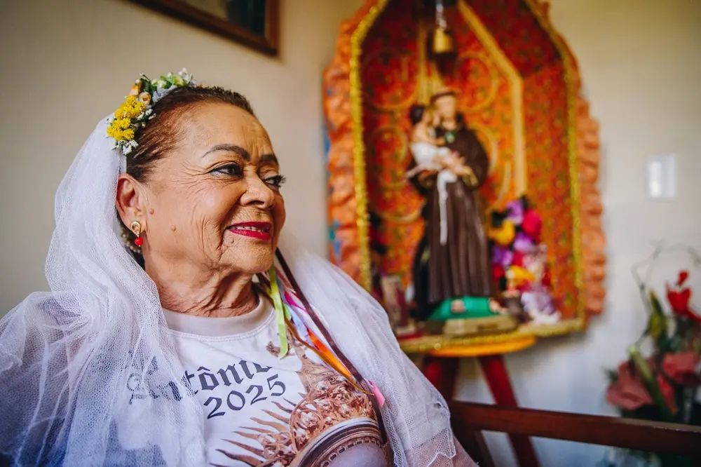 Na imagem, retrato em plano médio de uma mulher idosa sorridente, vista de perfil parcial, olhando para a direita. Ela tem pele madura, olhos maquiados e usa um batom vermelho vibrante. Em sua cabeça, há um véu de noiva feito de tule branco preso por uma tiara de flores amarelas e brancas, além de fitas coloridas pendendo na lateral. Ela veste uma camiseta branca estampada com uma imagem religiosa e o texto parcial 