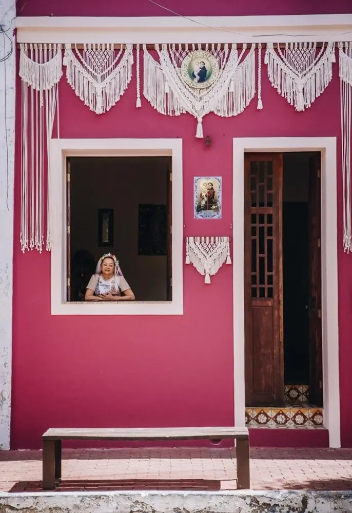 Na imagem, fotografia em plano médio de uma mulher idosa, identificada como Socorro Luna, olhando para cima com expressão de devoção. Ela usa um véu de noiva branco curto com arranjo de flores na cabeça e uma camiseta branca com a estampa 