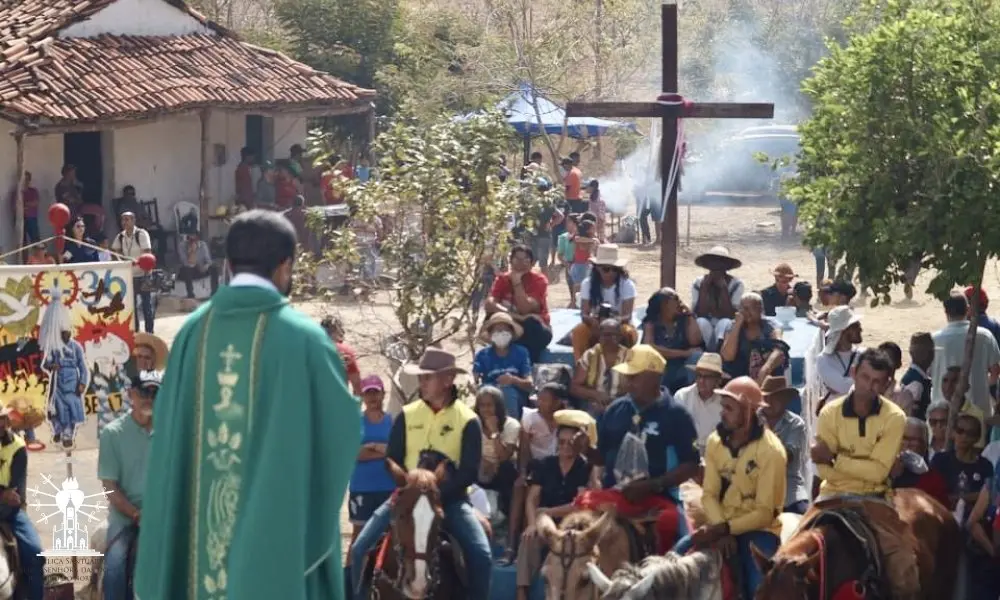 Fotografia tirada pelas costas de um padre de túnica verde, que celebra uma missa campal na imagem anderson araújo.jpeg. Diante dele, fiéis e vaqueiros montados a cavalo assistem à cerimônia em uma área rural. Ao fundo, há uma grande cruz de madeira fixa no chão, fumaça dispersa e uma casa simples com telhado de barro. O dia está ensolarado.