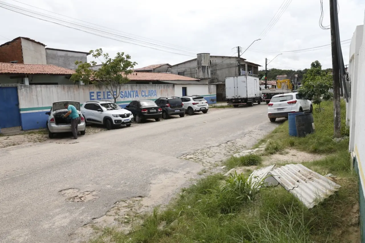 Carros estacionados em rua residencial em frente ao muro da escola E.E.I.E.F. Santa Clara. À esquerda, um homem mexe no porta-malas de um veículo.