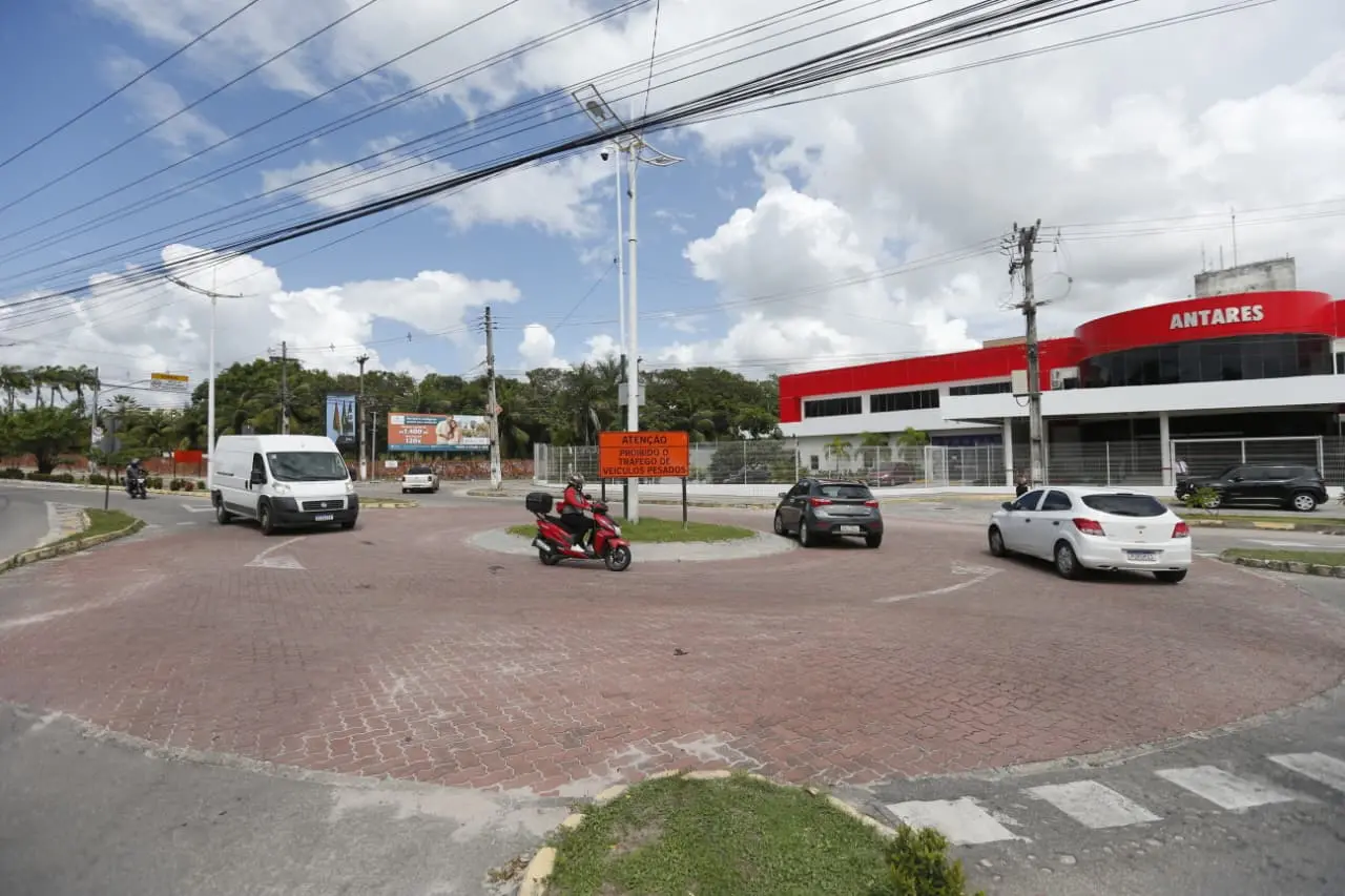 Fluxo de veículos em rotatória no Eusébio, Ceará. Carros, uma van branca e uma moto contornam a via em frente a um prédio comercial com fachada vermelha.