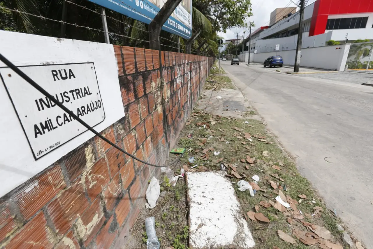 Calçada com lixo e folhas na Rua Industrial Amilcar Araújo, no Eusébio. Em destaque, placa da via fixada em muro de tijolos com fio pendurado.