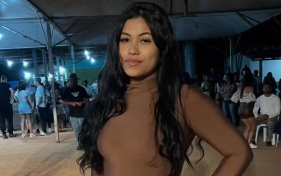 Geniane Pereira, jovem de cabelos longos e escuros posa para a foto em um evento noturno, usando blusa de gola alta em tom marrom. Ao fundo, há grupos de pessoas reunidas sob uma estrutura coberta, com iluminação artificial e ambiente de confraternização.