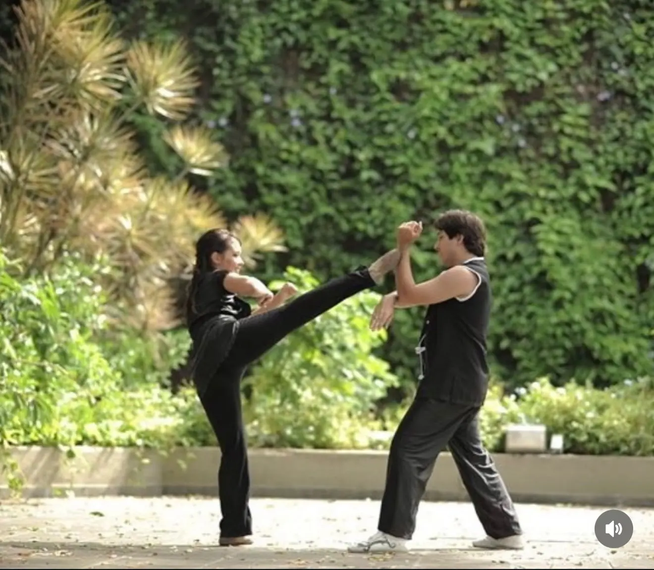 Na imagem, fotografia em plano inteiro mostra um homem e uma mulher praticando artes marciais em um espaço ao ar livre com piso de pedra. À esquerda, a mulher está de perfil, equilibrada em uma perna só, enquanto executa um chute alto lateral com a perna direita estendida em direção ao homem. Ela veste uma blusa e calça compridas, ambas pretas. À direita, o homem está posicionado de perfil para ela, em uma base firme com os joelhos levemente flexionados, usando os antebraços erguidos para bloquear o chute. Ele veste um traje tradicional de treino preto sem mangas com detalhes em branco e calças pretas folgadas. O cenário ao fundo é composto por uma parede densamente coberta por uma trepadeira verde e, à esquerda, arbustos e plantas ornamentais de tons esverdeados e amarelados sob a luz do dia. No canto inferior direito da imagem, há um pequeno ícone cinza de alto-falante.