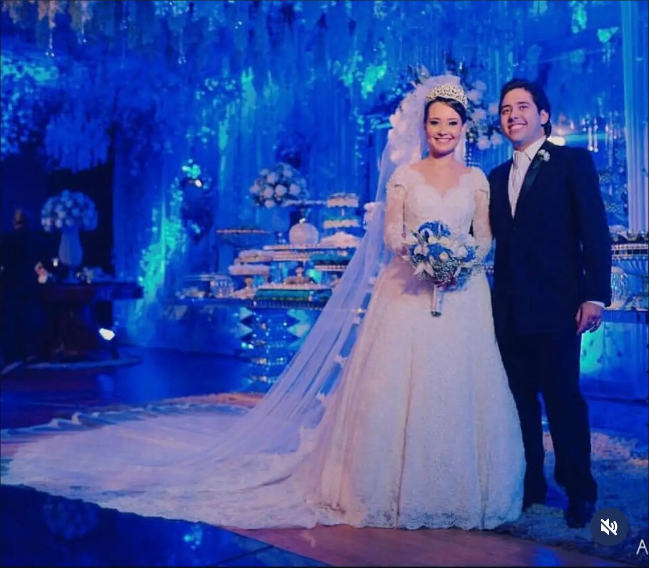 Na imagem, uma fotografia de plano inteiro mostra um casal de noivos sorridentes, em pé e lado a lado, durante a recepção de um casamento. O ambiente é luxuoso, decorado com uma iluminação cênica predominantemente azul vibrante. A noiva, à esquerda, ela usa um vestido de noiva clássico na cor branca, com mangas longas em renda e decote em 