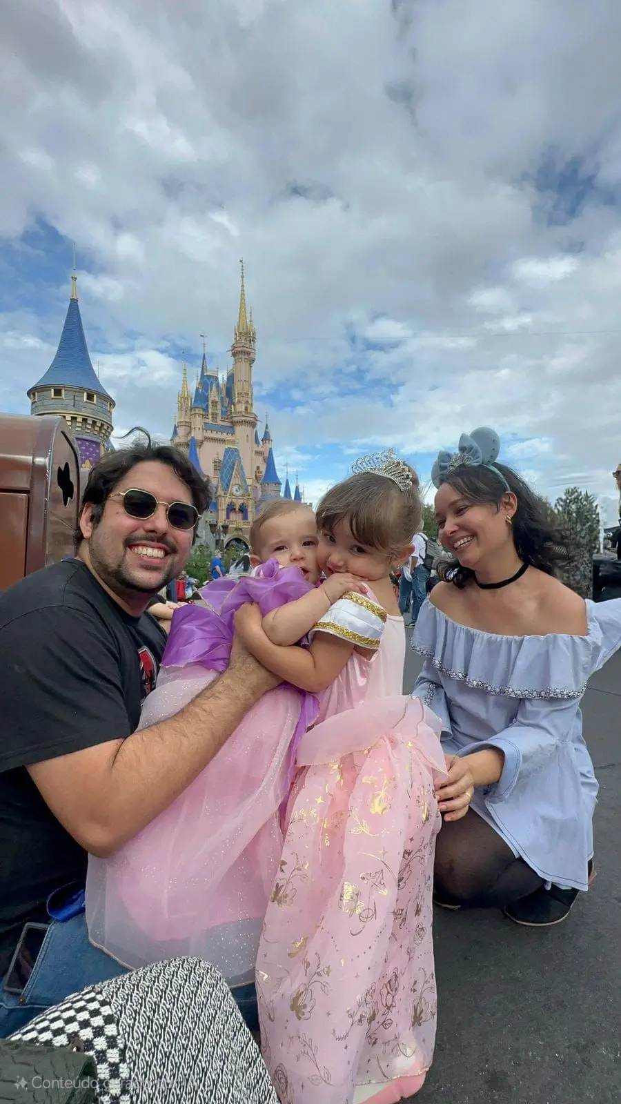 Na imagem, uma foto colorida ao ar livre mostra uma família de quatro pessoas sorrindo em frente ao Castelo da Cinderela, no Magic Kingdom, Disney. À esquerda, um homem de barba, óculos escuros e camiseta preta sorri para a câmera enquanto segura uma bebê vestida com um traje de princesa roxo. No centro, uma menina pequena com uma tiara de cristais e um vestido de princesa rosa abraça carinhosamente a bebê. À direita, uma mulher com tiara de orelhas do Mickey azuis e blusa azul claro de ombros de fora olha com ternura para as crianças. O céu está parcialmente nublado e o castelo icônico se destaca ao fundo em tons de azul e dourado.