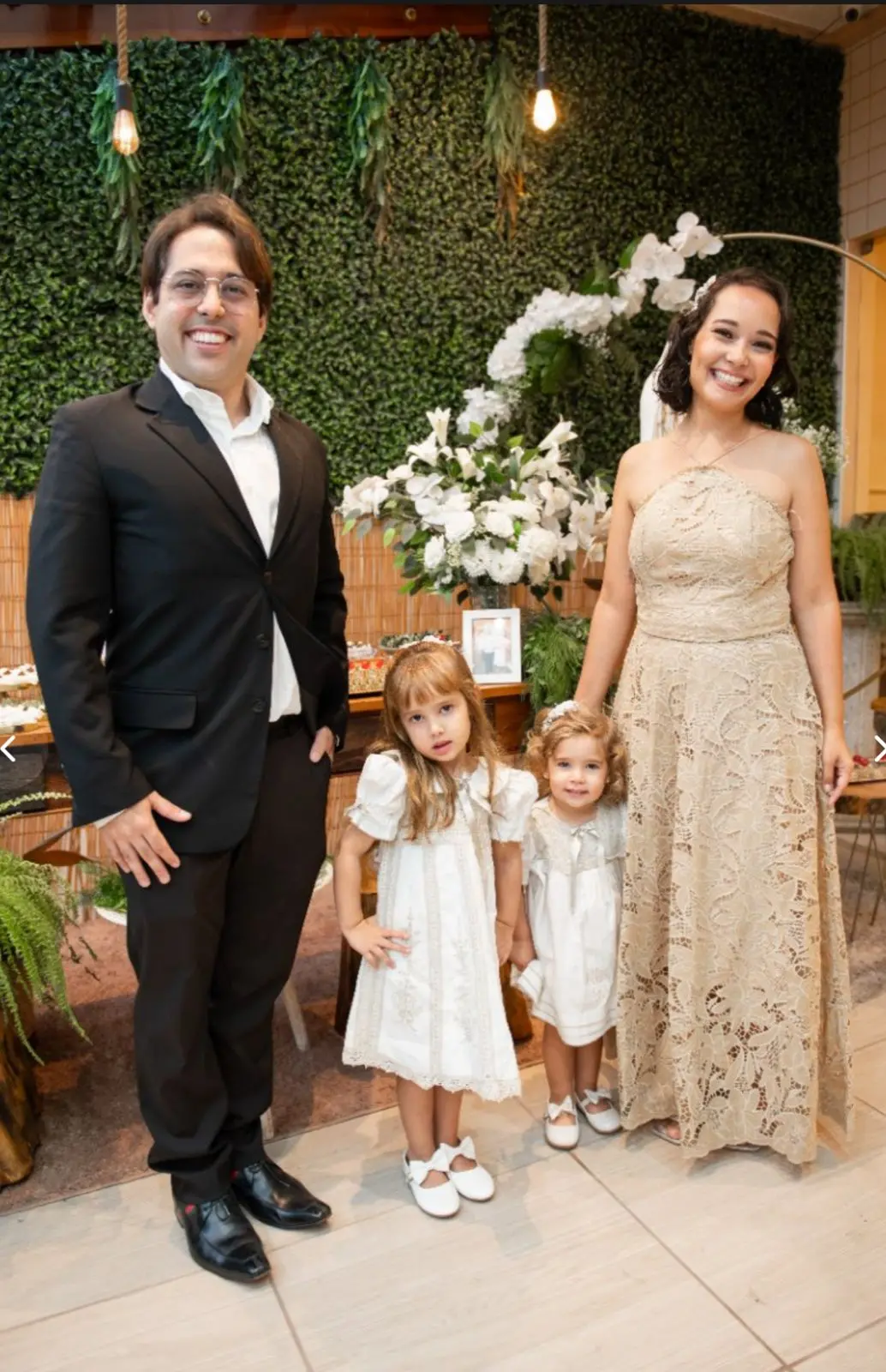 Na imagem, uma foto de plano médio mostra uma família de quatro pessoas posando sorridentes em um evento social. À esquerda, um homem usa óculos, paletó preto, camisa branca e sapatos sociais pretos. À direita, uma mulher sorri e usa um vestido longo de renda bege sem mangas. No centro, entre os adultos, estão duas meninas pequenas: a maior usa um vestido branco de mangas bufantes e a menor, com cabelos cacheados, usa um vestido branco similar e segura a mão da mulher. O fundo é decorado com um painel de folhagens verdes, arranjos de flores brancas sobre uma mesa de madeira e lâmpadas de filamento penduradas, criando um ambiente quente e elegante.