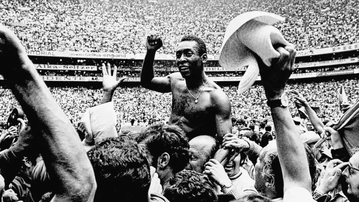 Fotografia em preto e branco mostra Pelé sendo carregado nos ombros por uma multidão dentro de um estádio lotado. Sem camisa, o jogador sorri e acena para o público enquanto torcedores levantam os braços ao seu redor em celebração. Ao fundo, as arquibancadas aparecem repletas de espectadores.