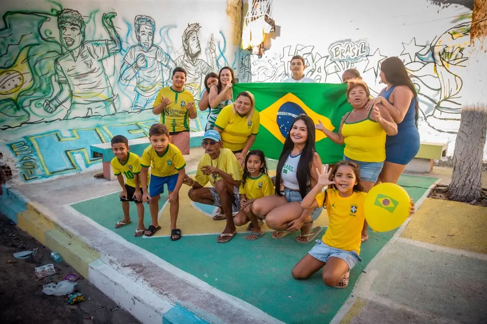 Na imagem, fotografia colorida ao ar livre de um grupo de cerca de doze pessoas de várias idades — crianças, jovens e adultos — reunidas e sorridentes em uma calçada ou praça pintada de verde e amarelo. A maioria delas veste camisas da seleção brasileira de futebol ou roupas nas cores amarela, verde e azul. No centro, algumas pessoas seguram aberta uma grande bandeira do Brasil. À direita, uma menina ajoelhada em primeiro plano segura um balão amarelo com a bandeira do Brasil estampada e acena para a câmera. Ao fundo, há um muro branco com grafites e desenhos estilizados de jogadores de futebol com traços em azul e verde, além da palavra 