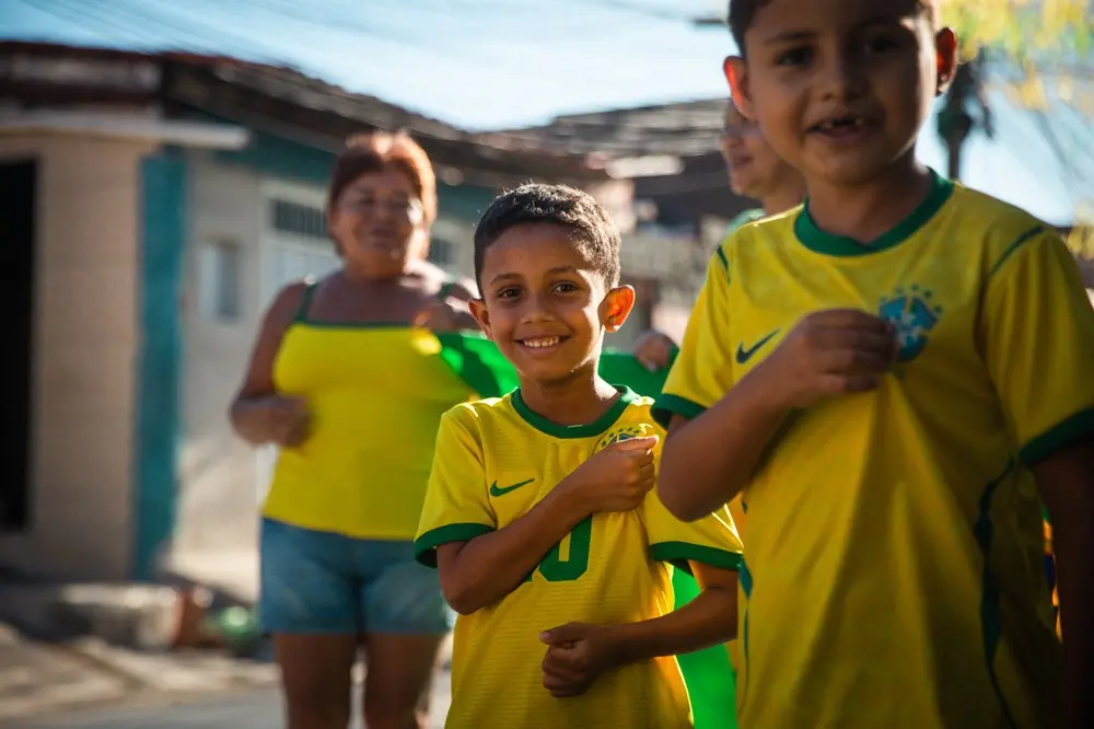 Na imagem, fotografia colorida em plano médio que destaca duas crianças vestindo a camisa amarela da seleção brasileira de futebol. No centro da imagem, um menino sorri diretamente para a câmera, com a mão esquerda próxima ao peito. À direita, em primeiro plano, aparece a metade do corpo de outro menino, ligeiramente desfocado, também usando a camisa da seleção. Ao fundo, com foco suave, uma mulher com blusa amarela e shorts jeans caminha segurando uma bandeira verde ao lado de outra pessoa. O cenário é uma rua residencial ao ar livre em um dia ensolarado, com casas simples ao fundo.