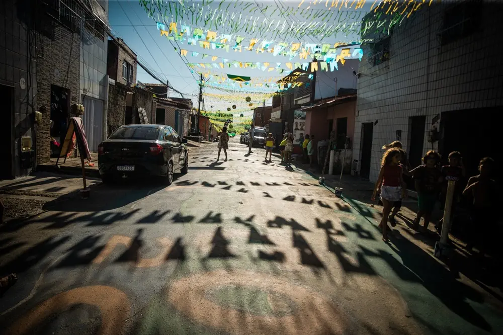 Na imagem, fotografia colorida ao nível da rua mostrando uma via residencial decorada para a Copa do Mundo sob a luz forte do sol. O asfalto está pintado com desenhos em tons de verde, amarelo e azul, e as sombras marcantes das bandeirinhas juninas penduradas no alto cobrem grande parte do chão em primeiro plano. À esquerda, um carro preto está estacionado de costas. Ao longo da rua, algumas pessoas caminham e conversam, e à direita, em primeiro plano, um grupo de crianças pequenas anda pela calçada na sombra. As casas nas laterais variam entre fachadas de tijolo e azulejos claros, sob um céu azul e limpo.