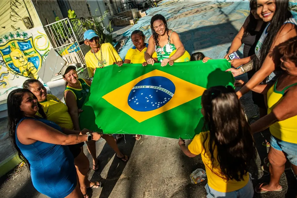 Na imagem, fotografia colorida com ângulo levemente inclinado (plongée) mostrando um grupo de cerca de dez pessoas reunidas em círculo em uma rua, segurando juntas uma grande bandeira do Brasil aberta horizontalmente no centro. O grupo é composto por homens, mulheres e crianças sorridentes, a maioria vestindo roupas em tons de amarelo, verde e azul. À esquerda, em primeiro plano, uma mulher de vestido azul sorri olhando para o grupo; à direita, uma menina de costas com blusa amarela segura uma das pontas da bandeira. Ao fundo, o asfalto da rua exibe pinturas em tons de azul e amarelo, e um muro à esquerda traz um grafite estilizado com o brasão da seleção brasileira e o desenho do 