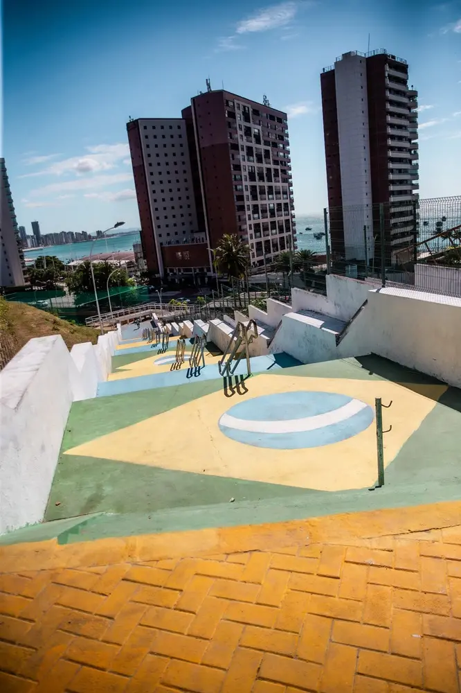 Na imagem, vista de cima de uma grande escadaria pública descendente. Os patamares planos entre os lances de degraus são decorados com a pintura estilizada da bandeira brasileira, exibindo o losango amarelo sobre o fundo verde e o círculo azul cortado por uma faixa branca. A escadaria possui corrimãos centrais de metal e é ladeada por paredes brancas escalonadas. Na base da colina, a paisagem urbana se revela com prédios altos de arquitetura moderna e, ao longe, a linha costeira com o mar azul e embarcações distantes sob um céu ensolarado.