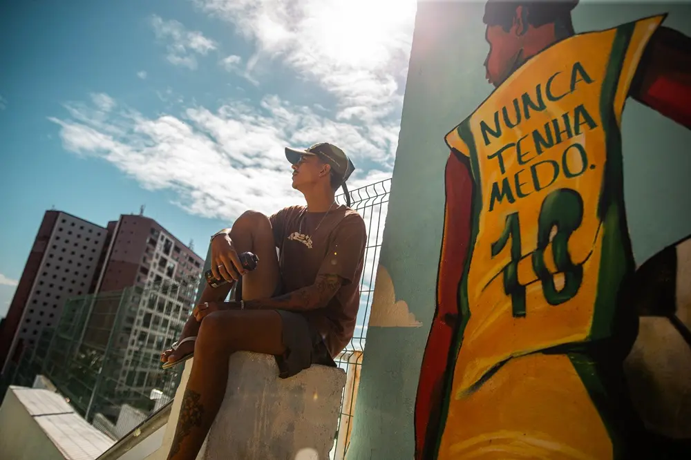 Na imagem, uma fotografia tirada de um ângulo baixo mostra um jovem tatuado, sentado em uma estrutura de concreto alta, com uma vista urbana ao fundo. Ele usa um boné verde e marrom, óculos, uma camiseta marrom e shorts, e segura uma lata de spray na mão direita. Ele olha para cima e para a direita, com o sol brilhando intensamente acima dele. À direita, um grande mural colorido em uma parede de concreto retrata as costas de um jogador de futebol vestindo uma camisa amarela e verde, com as palavras 