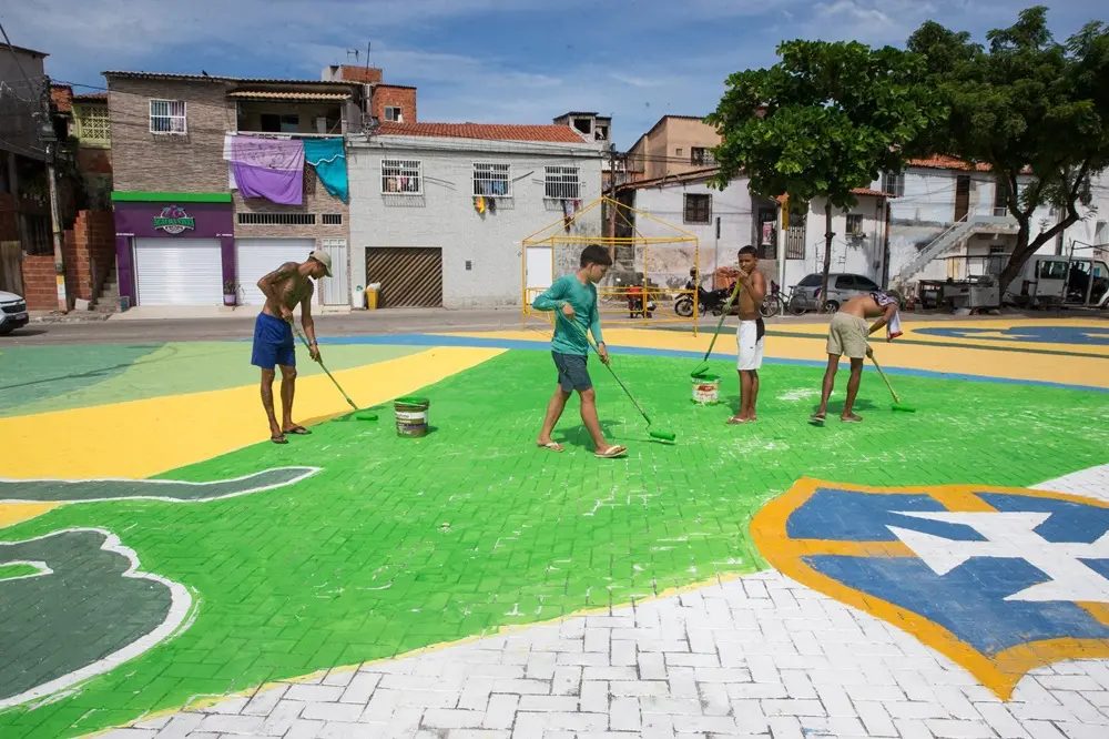 Na imagem, fotografia colorida em plano médio captura quatro jovens pintando o asfalto em uma praça urbana no bairro Castelo Encantado, em Fortaleza. O chão está pintado com o brasão da Confederação Brasileira de Futebol (CBF) nas cores verde, amarela e branca, em um design para a Copa do Mundo. À esquerda, um jovem de bermuda azul e sem camisa usa um rolo com cabo longo para aplicar tinta verde clara em uma grande seção do asfalto. No centro, um jovem de camiseta verde clara e bermuda escura usa outro rolo com cabo longo. Mais à direita, dois jovens pintam seções menores: um de bermuda branca e sem camisa usa um rolo mais curto, e um de bermuda bege e sem camisa pinta um detalhe em uma borda branca. Baldes de tinta verdes e brancos estão próximos a eles no chão. O fundo é composto por prédios residenciais e comerciais de alvenaria, com fachadas brancas, cinzas e marrons. À esquerda, destaca-se uma loja com uma placa roxa onde se lê 