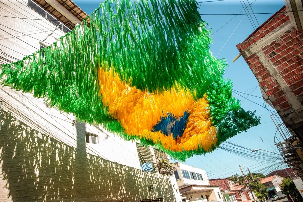 Na imagem, fotografia colorida tirada de baixo para cima (ângulo contra-plongé) destaca uma decoração aérea de Copa do Mundo em uma rua residencial. Centenas de fitas ou franjas de plástico fino estão amarradas em fios dispostos de um lado ao outro da rua, formando o desenho estilizado da bandeira do Brasil no ar. O topo e as laterais da composição são formados por franjas verdes, o centro traz um losango feito com franjas amarelas e, no meio, há um círculo de franjas azuis e brancas. A luz solar passa entre os plásticos, tornando as cores vibrantes. As franjas estão estendidas entre os prédios da vizinhança. À esquerda, vê-se a fachada texturizada de uma casa de dois andares de cor clara, com a sombra da decoração projetada sobre a parede. À direita, aparece parte de uma casa de tijolos ecológicos ou cerâmicos expostos, sem reboco. Cabos de energia cruzam o céu azul e limpo ao fundo, completando o cenário urbano da comunidade.