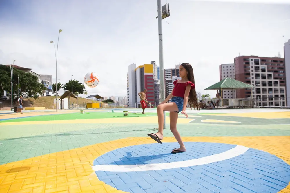 Na imagem, fotografia colorida em plano médio e ângulo levemente baixo captura uma menina brincando com uma bola em um espaço público pavimentado com blocos de concreto intertravados. O chão da praça está pintado em faixas horizontais de verde e amarelo, e a menina está posicionada exatamente sobre um grande círculo azul cortado por uma faixa branca, reproduzindo o centro da bandeira do Brasil. A menina tem cabelos longos e castanhos, veste uma blusa vermelha de mangas curtas no estilo ciganinha, short jeans curto e chinelos de dedo escuros. Ela está com a perna direita suspensa e flexionada no ar, equilibrando-se no pé esquerdo em pleno movimento para chutar ou controlar uma bola de futebol (branca com detalhes laranjas e pretos) que flutua à sua esquerda.
