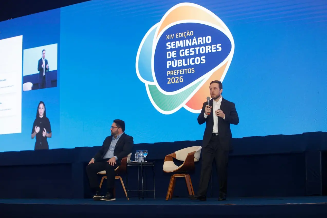 Diretor da Quaest faz palestra no palco do XIV Seminário dos Gestores Públicos – Prefeitos 2026. Em pé, ele segura um microfone diante de um telão com a identidade visual do evento, enquanto outro participante permanece sentado ao lado. Uma intérprete de Libras aparece em destaque na tela lateral.