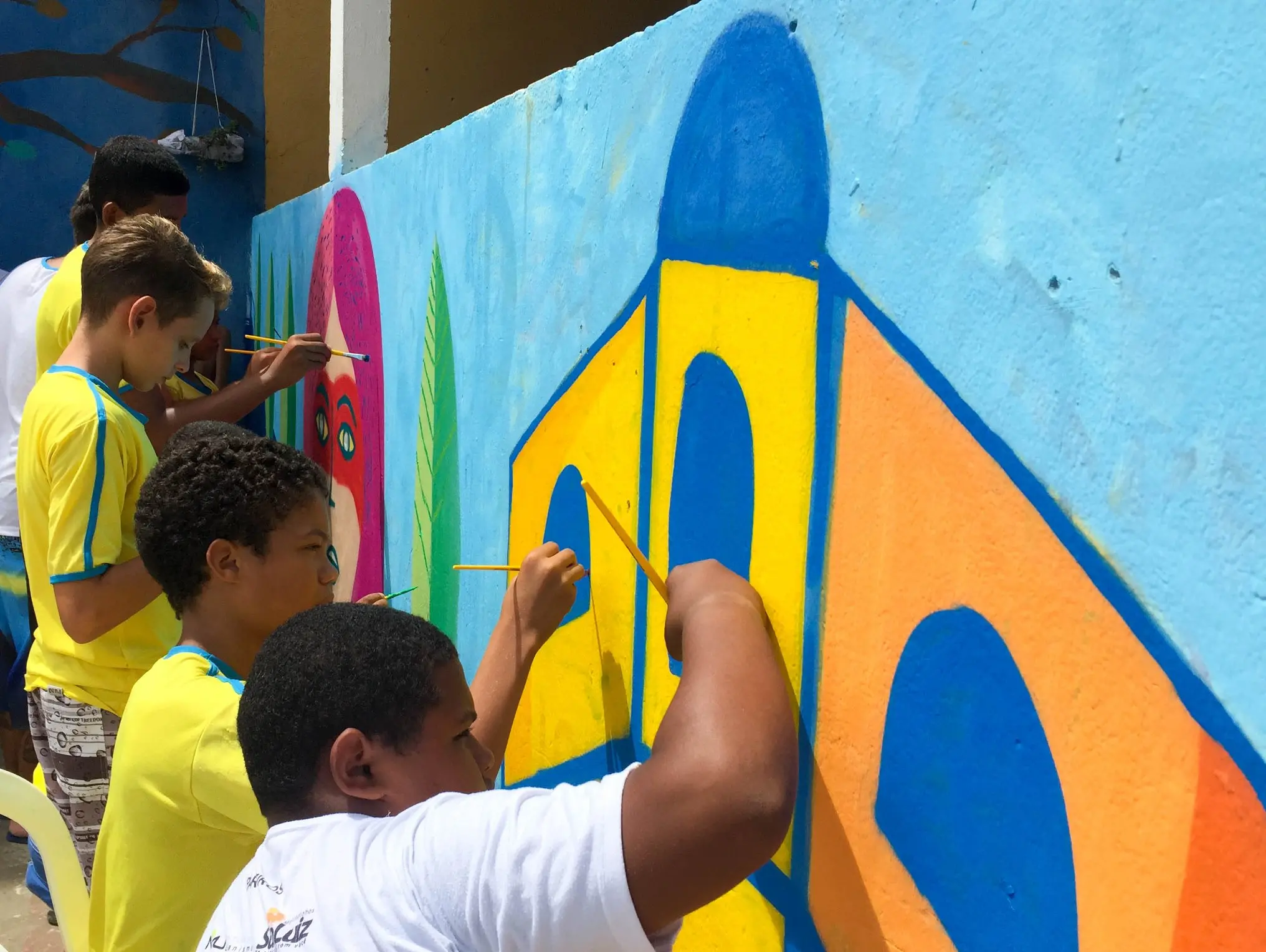 Estudantes vestindo camisas amarelas pintam um mural colorido em uma parede externa, como visto em Programa Escolas de Cultura do Ceará.