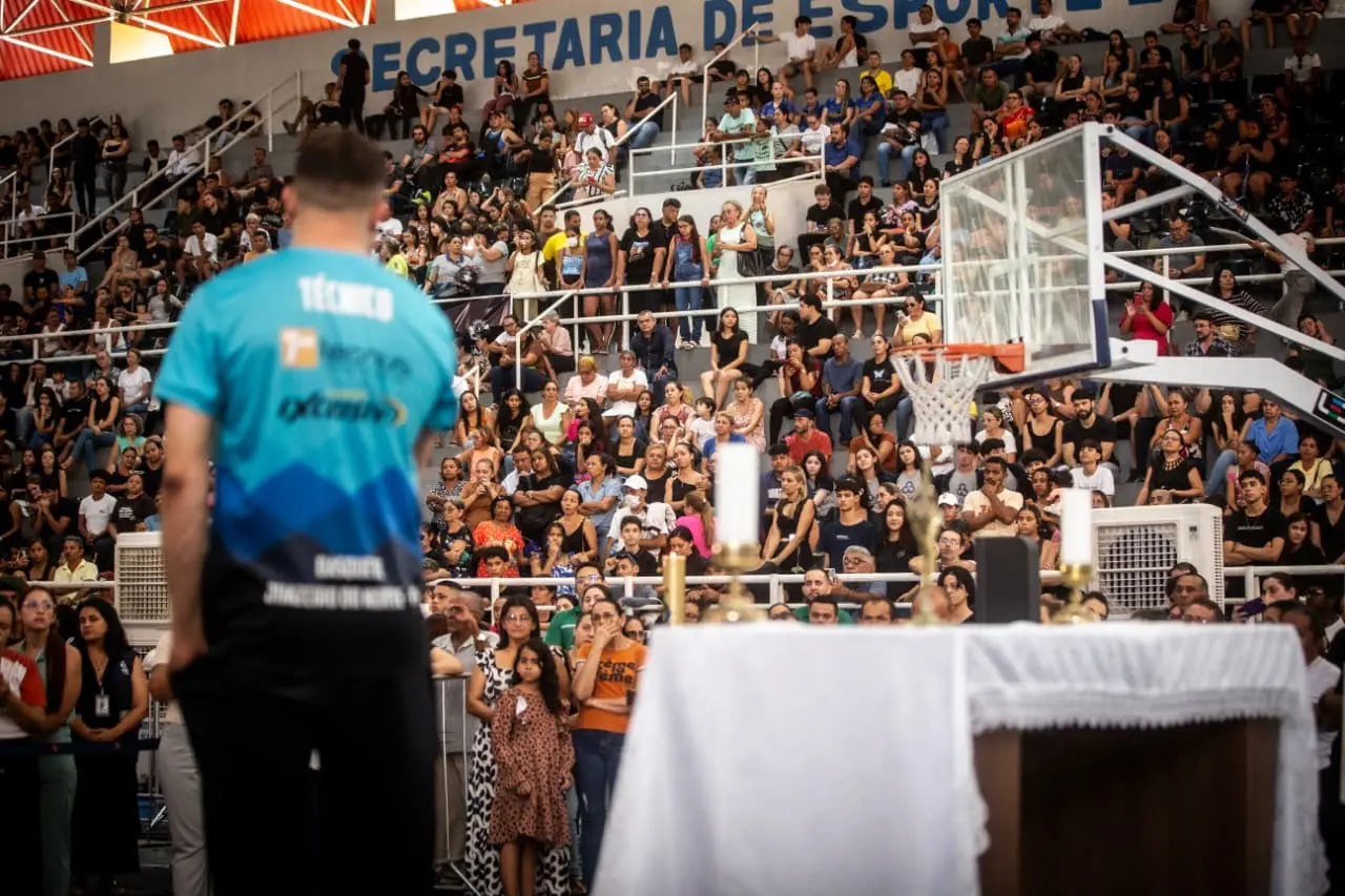 Foto mostra ginásio com arquibancadas lotadas. Um homem de costas, com blusa azul escrito 