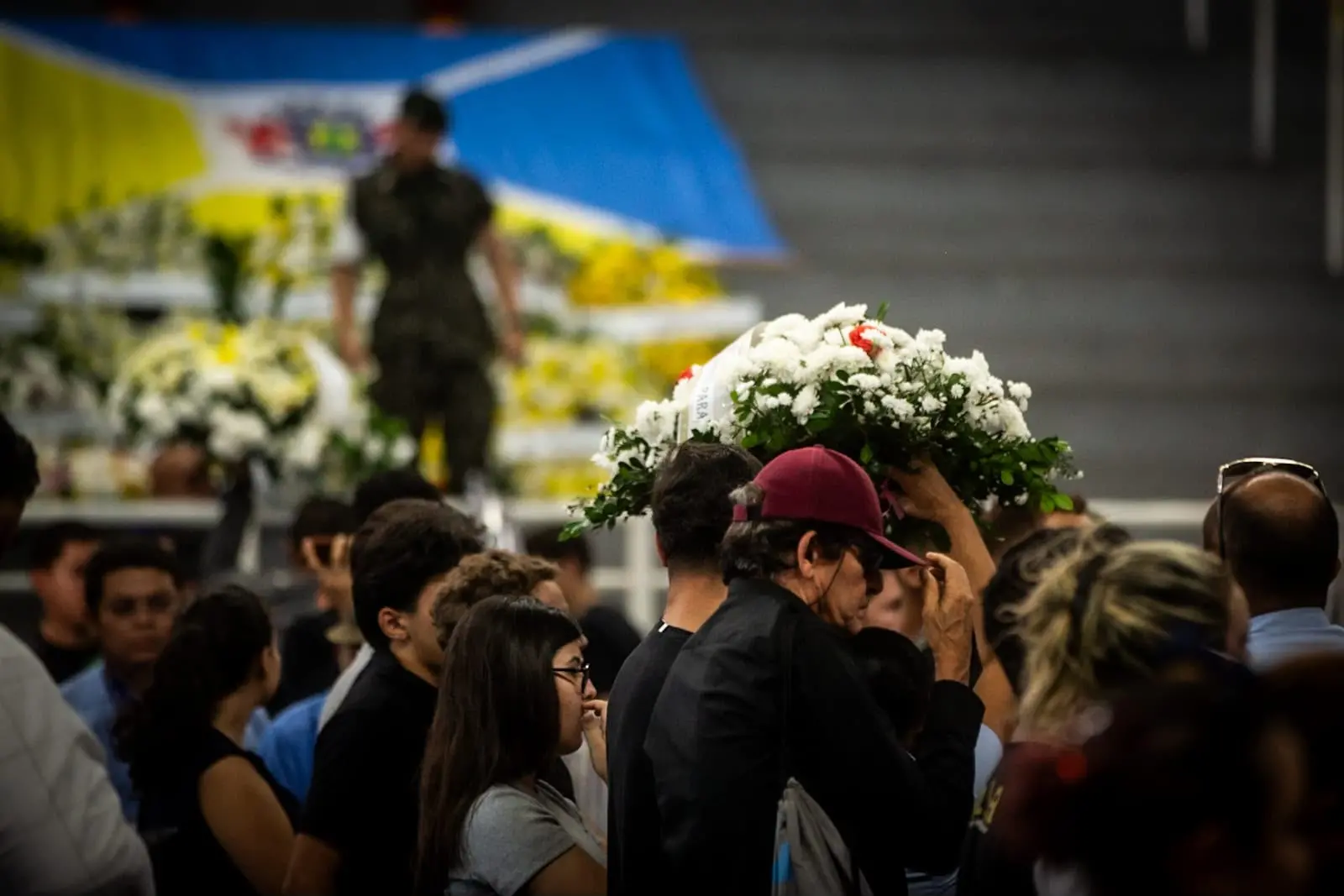 Foto mostra, em primeiro plano, pessoas carregando uma coroa de flores brancas. Ao fundo, a bandeira de Juazeiro do Norte está estendida na arquibancada do ginásio.