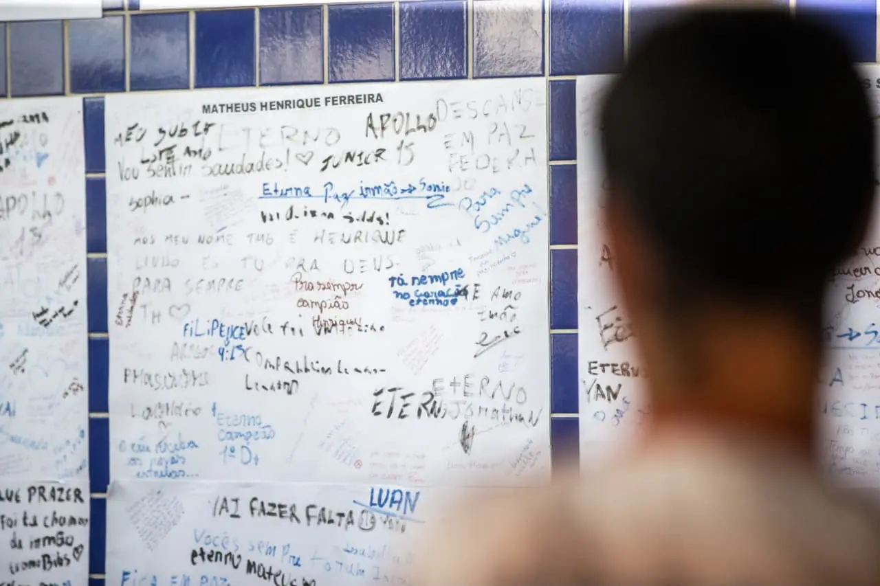 Na imagem, fotografia em plano médio com foco seletivo. Em primeiro plano, à direita, vê-se a nuca e os ombros desfocados de uma pessoa de cabelos curtos, de costas para a câmera. O foco da imagem está ao fundo, em uma parede revestida de azulejos azuis onde há cartazes brancos colados. Os cartazes estão cobertos de mensagens escritas à mão com canetas de várias cores, incluindo frases de luto e homenagem como 
