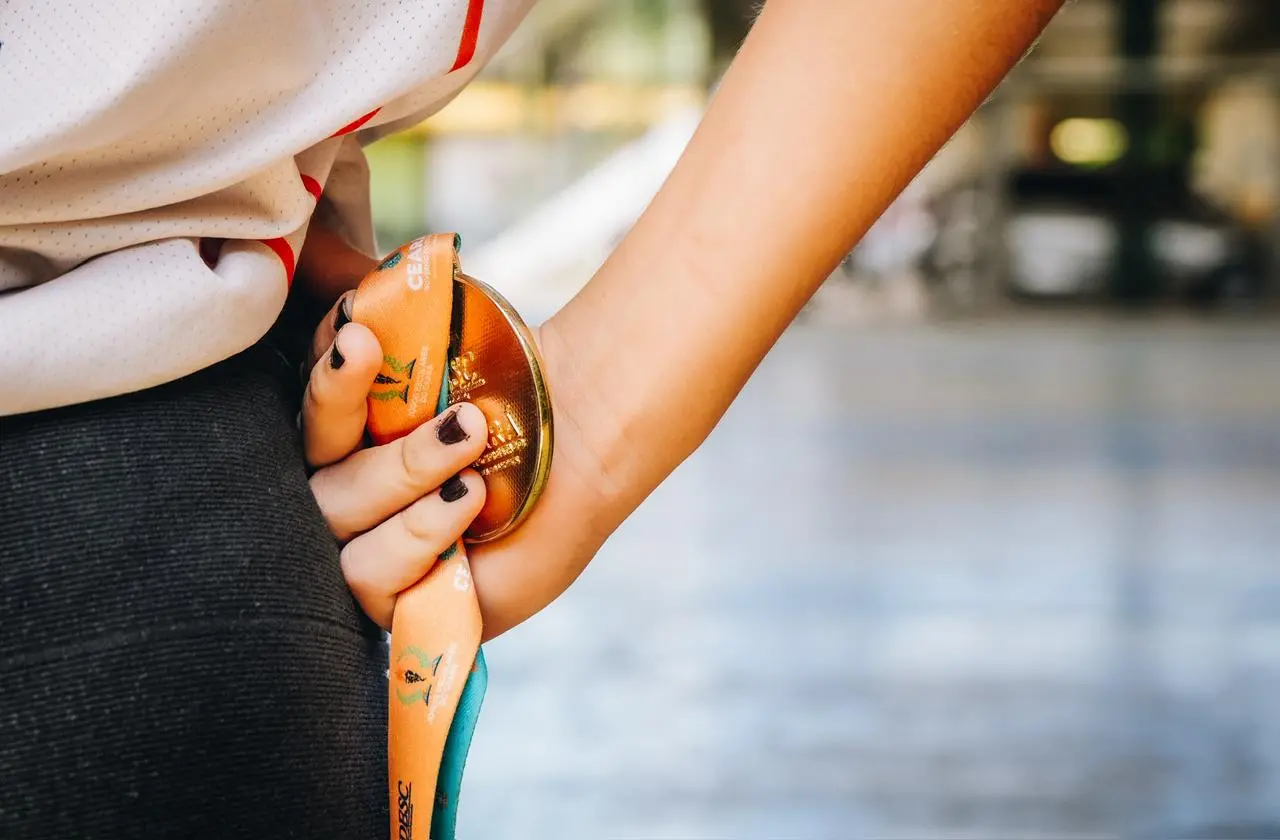 Na imagem, um close em cores foca na mão direita de uma pessoa, segurando uma medalha de ouro nas costas, logo abaixo da linha da cintura. A pessoa está usando uma camiseta branca esportiva com detalhes em vermelho e cinza, que é vista de costas, e uma calça escura. Seus dedos, com as unhas pintadas de preto, seguram a medalha dourada brilhante e sua fita laranja. A fita tem marcas sutis e letras, incluindo 