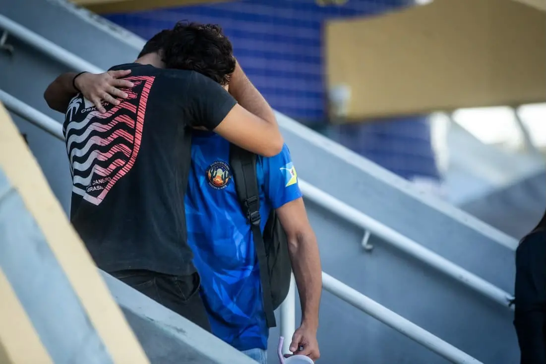 Na imagem, fotografia em plano médio capturada em ângulo diagonal. Duas pessoas se abraçam de forma consoladora nos degraus de concreto cinza de uma arquibancada. À esquerda, uma pessoa de costas veste uma camiseta preta com uma estampa geométrica ondulada em branco e vermelho que lembra o calçadão de Copacabana. Ela está com o braço esquerdo estendido sobre o ombro da outra pessoa, que veste uma camiseta azul-escura com detalhes geométricos mais claros e um brasão circular na manga. Ao fundo, detalhes em azul e amarelo completam a estrutura da arquibancada sob iluminação natural.