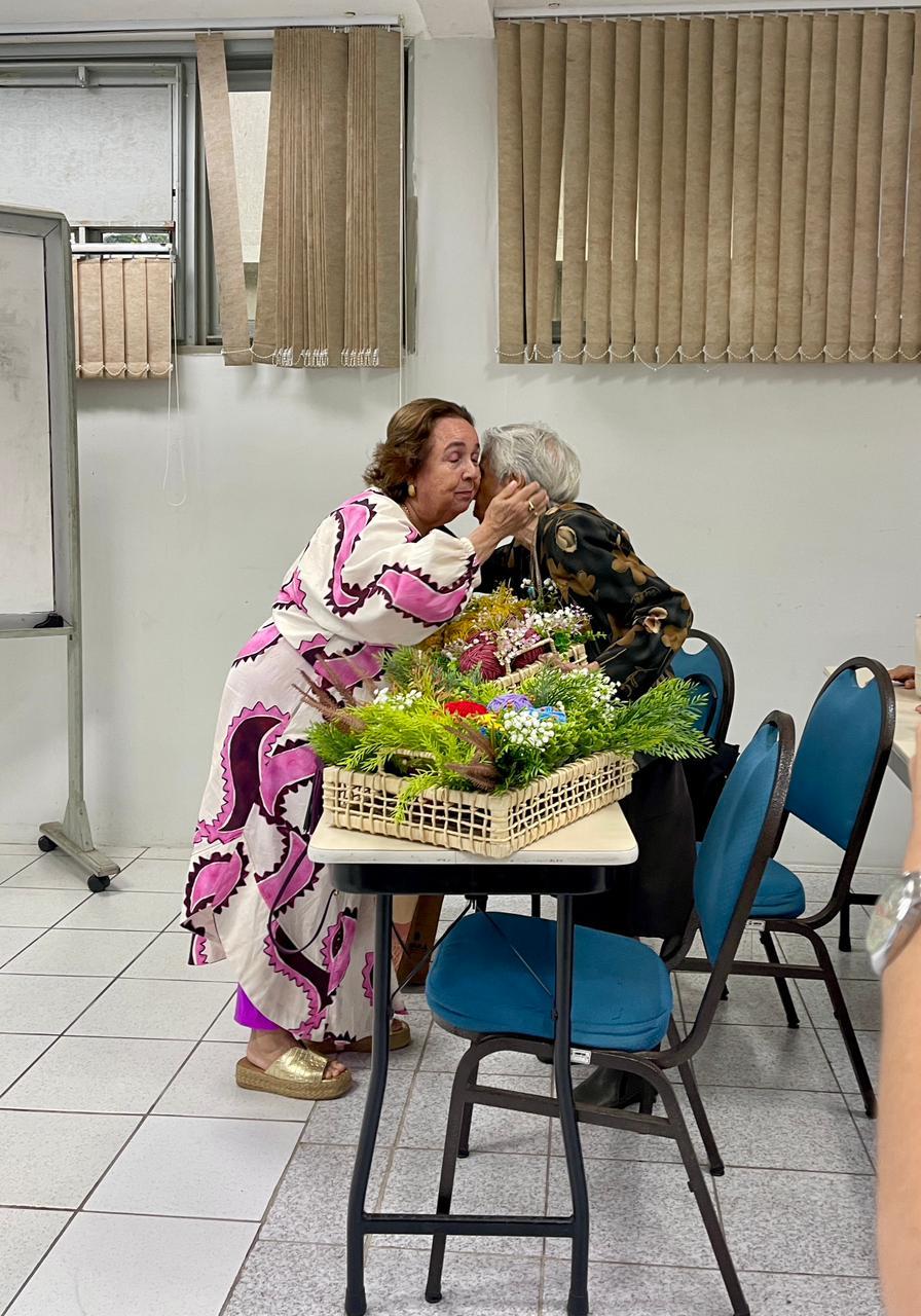 Na imagem, fotografia na vertical, em plano inteiro e de perfil, em uma sala de aula. À esquerda, uma mulher idosa de cabelos castanhos curtos (Dona Marivan) usa um vestido longo off-white com estampas abstratas em tons de rosa e roxo e sandálias de plataforma douradas. Ela está levemente inclinada para a frente, acariciando com carinho o rosto e dando um beijo na bochecha de outra mulher idosa de cabelos brancos curtos (Dona Maria Augusta). A segunda senhora, à direita, está sentada de costas para a câmera, vestindo uma camisa preta com estampa floral em tons de bronze. Entre elas, sobre uma mesa escolar de tampo claro e pernas pretas, há duas grandes cestas de palha trançada repletas de arranjos verdes, flores brancas miúdas e novelos de lã coloridos. Ao redor da mesa, aparecem cadeiras de metal com assento e encosto estofados em azul escuro. Ao fundo, a sala possui piso de lajotas claras, paredes brancas, uma lousa branca com rodinhas à esquerda e janelas altas cobertas por persianas verticais de tecido bege.