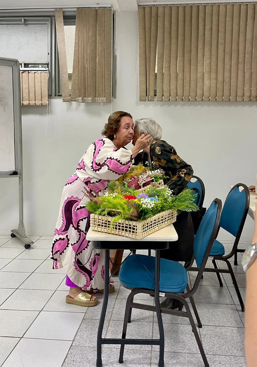 Na imagem, fotografia na vertical, em plano inteiro e de perfil, em uma sala de aula. À esquerda, uma mulher idosa de cabelos castanhos curtos (Dona Marivan) usa um vestido longo off-white com estampas abstratas em tons de rosa e roxo e sandálias de plataforma douradas. Ela está levemente inclinada para a frente, acariciando com carinho o rosto e dando um beijo na bochecha de outra mulher idosa de cabelos brancos curtos (Dona Maria Augusta). A segunda senhora, à direita, está sentada de costas para a câmera, vestindo uma camisa preta com estampa floral em tons de bronze. Entre elas, sobre uma mesa escolar de tampo claro e pernas pretas, há duas grandes cestas de palha trançada repletas de arranjos verdes, flores brancas miúdas e novelos de lã coloridos. Ao redor da mesa, aparecem cadeiras de metal com assento e encosto estofados em azul escuro. Ao fundo, a sala possui piso de lajotas claras, paredes brancas, uma lousa branca com rodinhas à esquerda e janelas altas cobertas por persianas verticais de tecido bege.