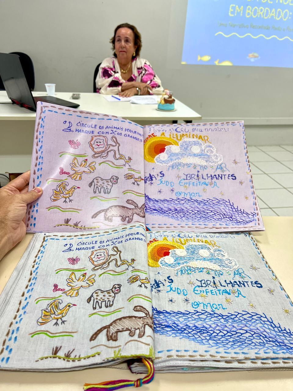 Na imagem, fotografia na vertical, em plano médio, com foco em dois livros abertos em primeiro plano sobre uma mesa clara. O livro de cima é segurado por uma mão no canto esquerdo e possui páginas em um tom lilás claro. O livro de baixo está apoiado na mesa e tem páginas em tecido cinza claro. Ambos exibem páginas duplas com o mesmo design pedagógico e poético bordado à mão: a página esquerda contém ilustrações bordadas de animais (leão, pássaros, ovelha, cachorro) e o texto