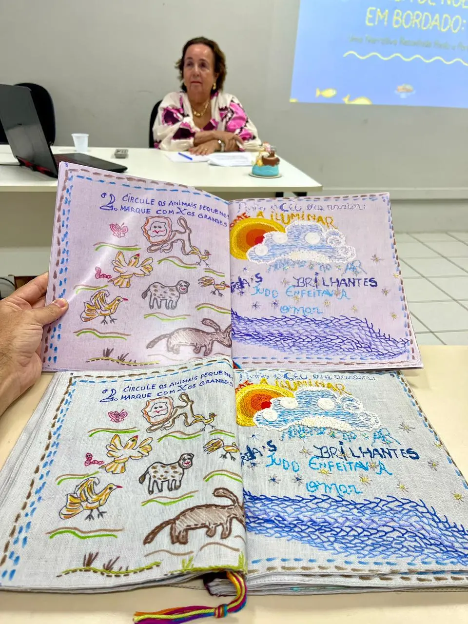 Na imagem, fotografia na vertical, em plano médio, com foco em dois livros abertos em primeiro plano sobre uma mesa clara. O livro de cima é segurado por uma mão no canto esquerdo e possui páginas em um tom lilás claro. O livro de baixo está apoiado na mesa e tem páginas em tecido cinza claro. Ambos exibem páginas duplas com o mesmo design pedagógico e poético bordado à mão: a página esquerda contém ilustrações bordadas de animais (leão, pássaros, ovelha, cachorro) e o texto 