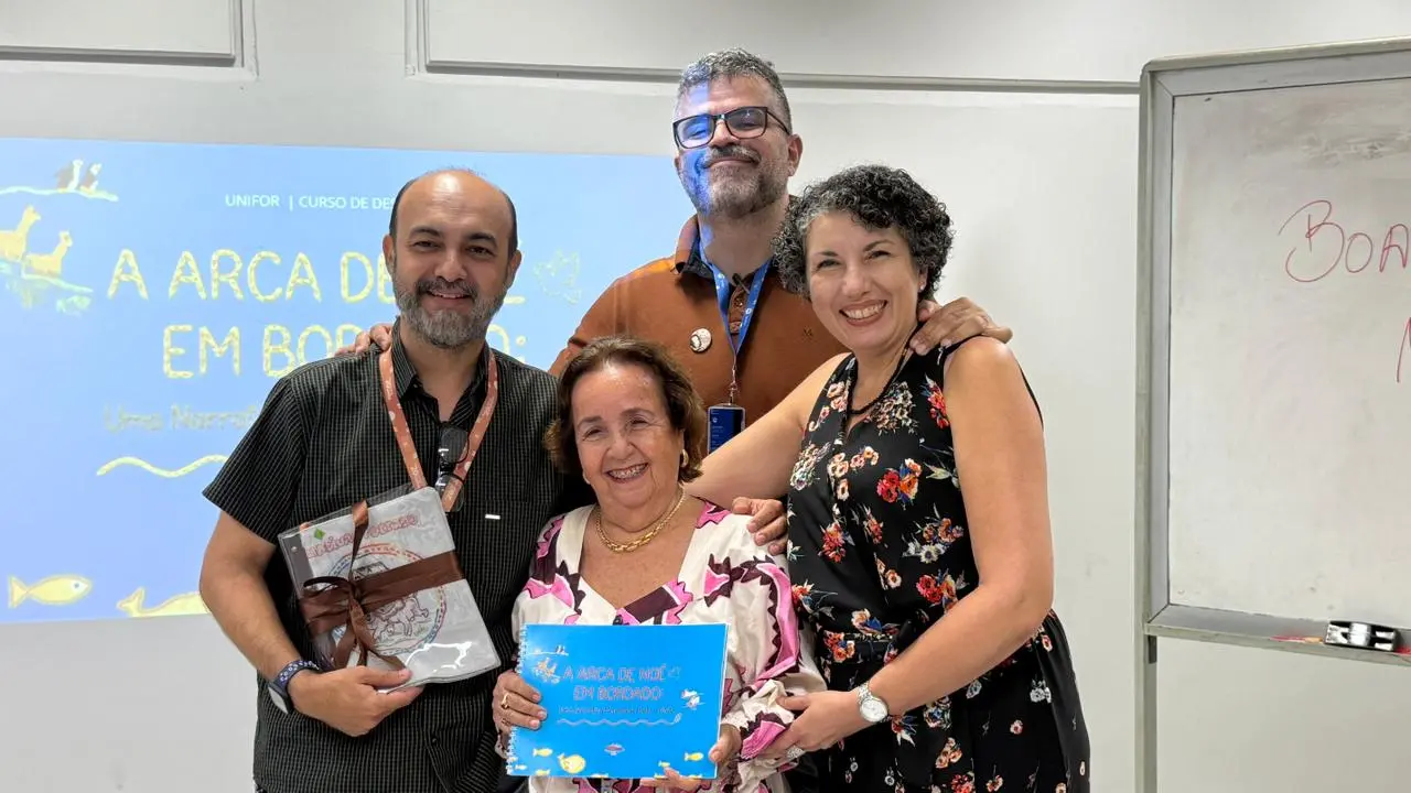 Na imagem, uma senhora de idade avançada, branca, com vestido florido e papel na mão, espécie de certificado, está abraçada com outras três pessoas. Um deles é careca e veste uma camisa preta; o que ocupa a posição central veste uma camisa polo bege; a pessoa que ocupa o lado direito é uma mulher com vestido preto estampado. O ambiente é claro e com luz branca fluorescente. Ao fundo, há uma imagem de projetor.